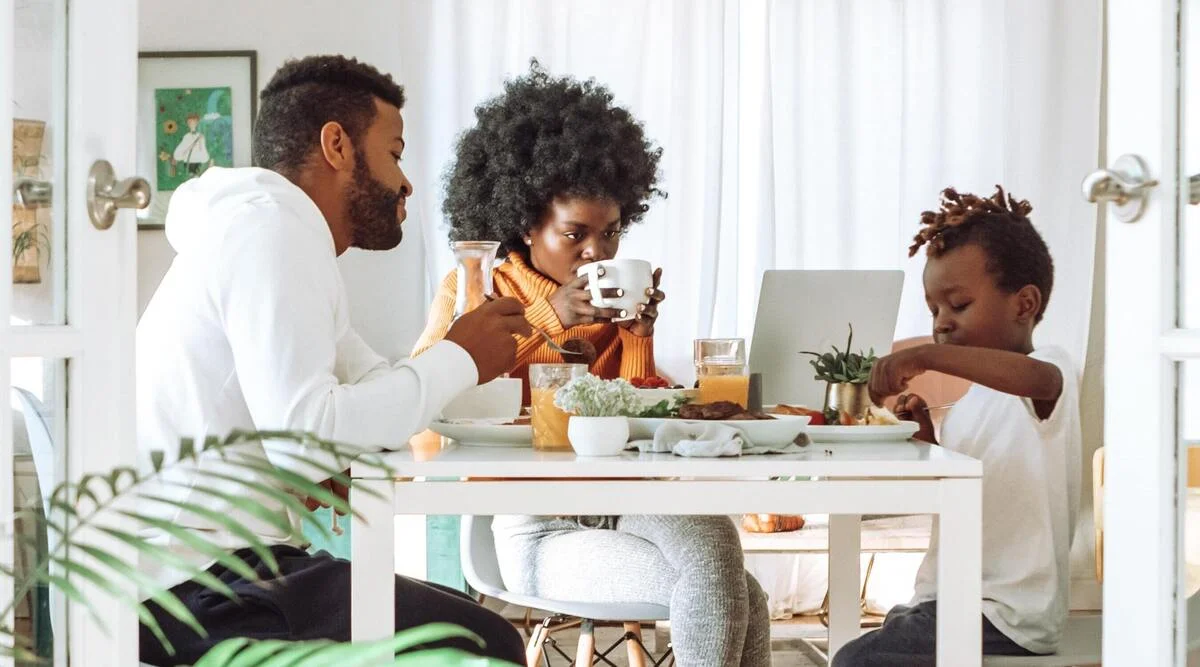 Parents and their son eating breakfast together at a bright kitchen table, a laptop open between them